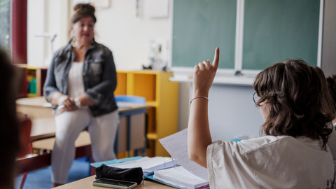 Im Klassenraum: Ein Schüler meldet sich. Im Hintergrund sitzt eine Pädagogin.