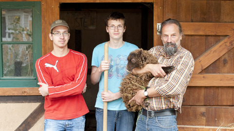 2 Schüler und 1 Pädagoge stehen vor dem Schafstall. Der Mann hält ein kleines, braunes Schaf auf dem Arm.