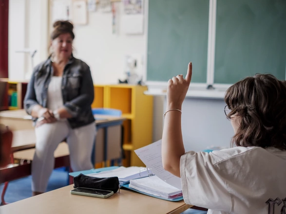 Im Klassenraum: Ein Schüler meldet sich. Im Hintergrund sitzt eine Pädagogin.