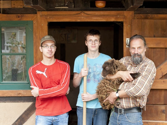 2 Schüler und 1 Pädagoge stehen vor dem Schafstall. Der Mann hält ein kleines, braunes Schaf auf dem Arm.